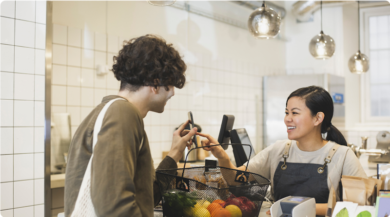 A young man uses mobile pay to purchase the produce in his shopping basket. The cashier smiles as she touches his phone.