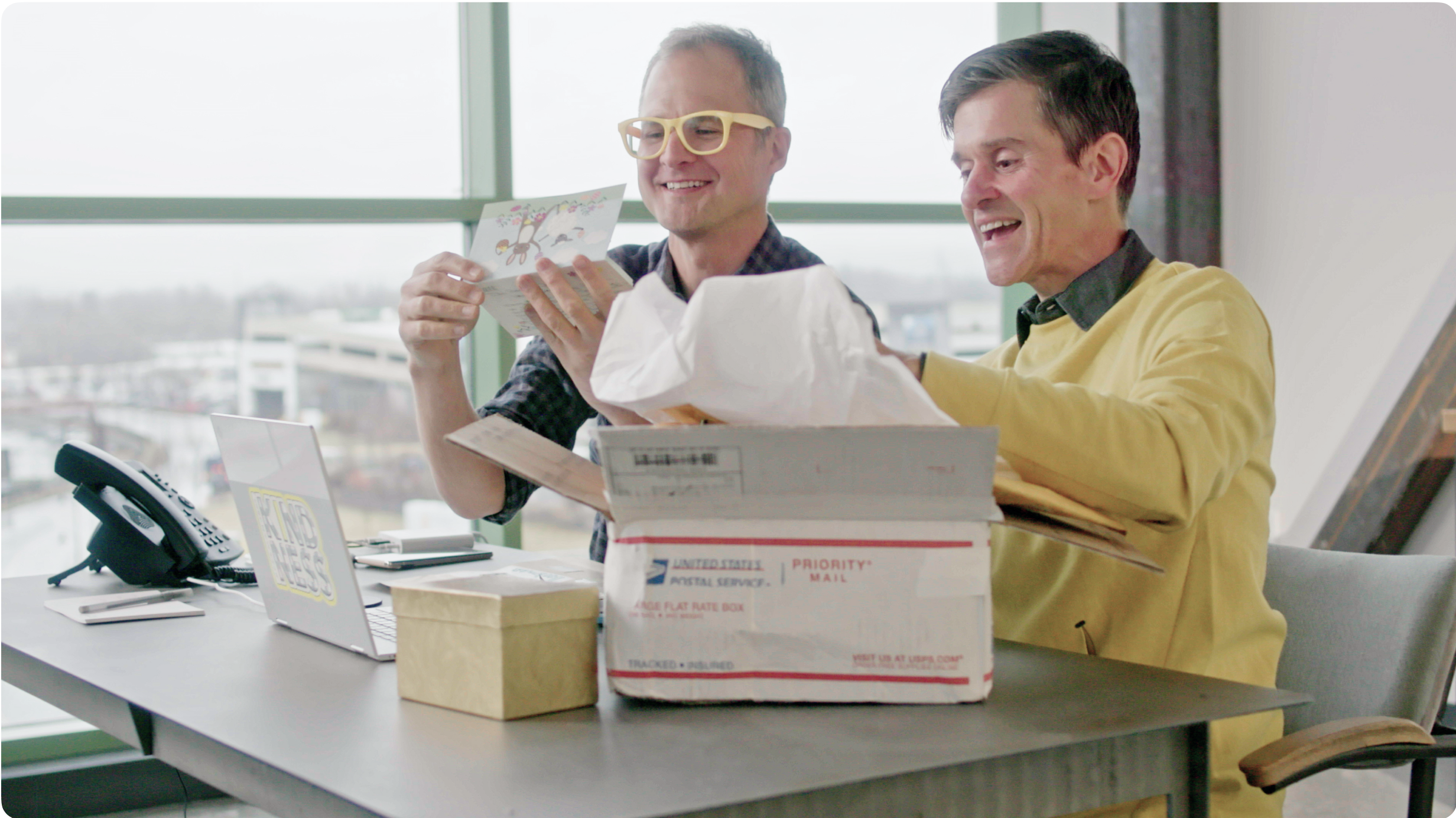 Josh and Brent open a package while sitting next to each other at a desk.