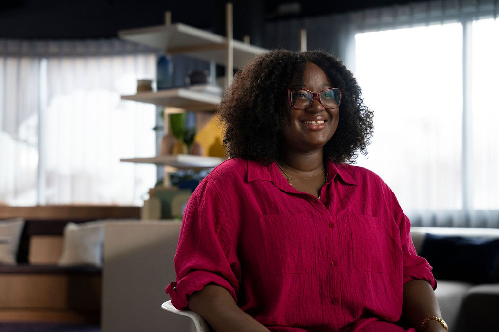 A woman with glasses in a red shirt smiles and speaks animatedly to camera against a blurred background.