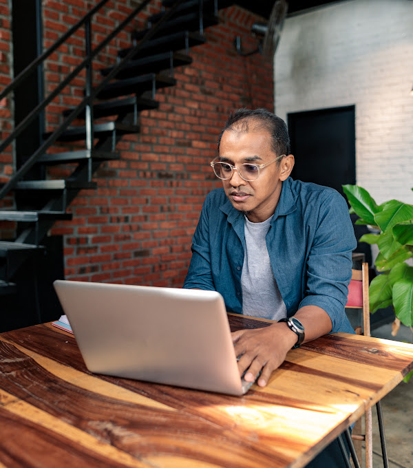 A man with glasses sits at a high top wooden table while working on his laptop.