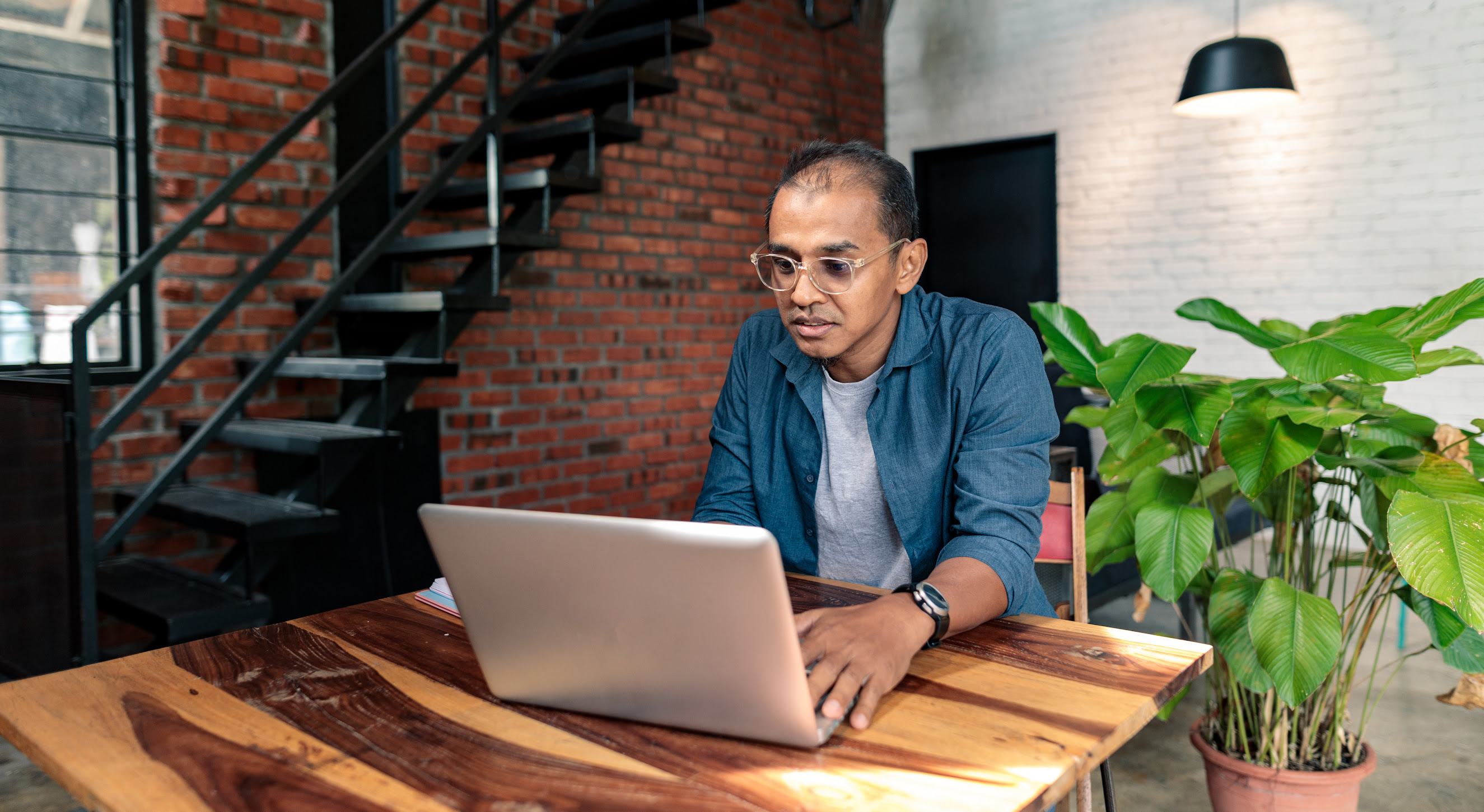 A man with glasses sits at a high top wooden table while working on his laptop.