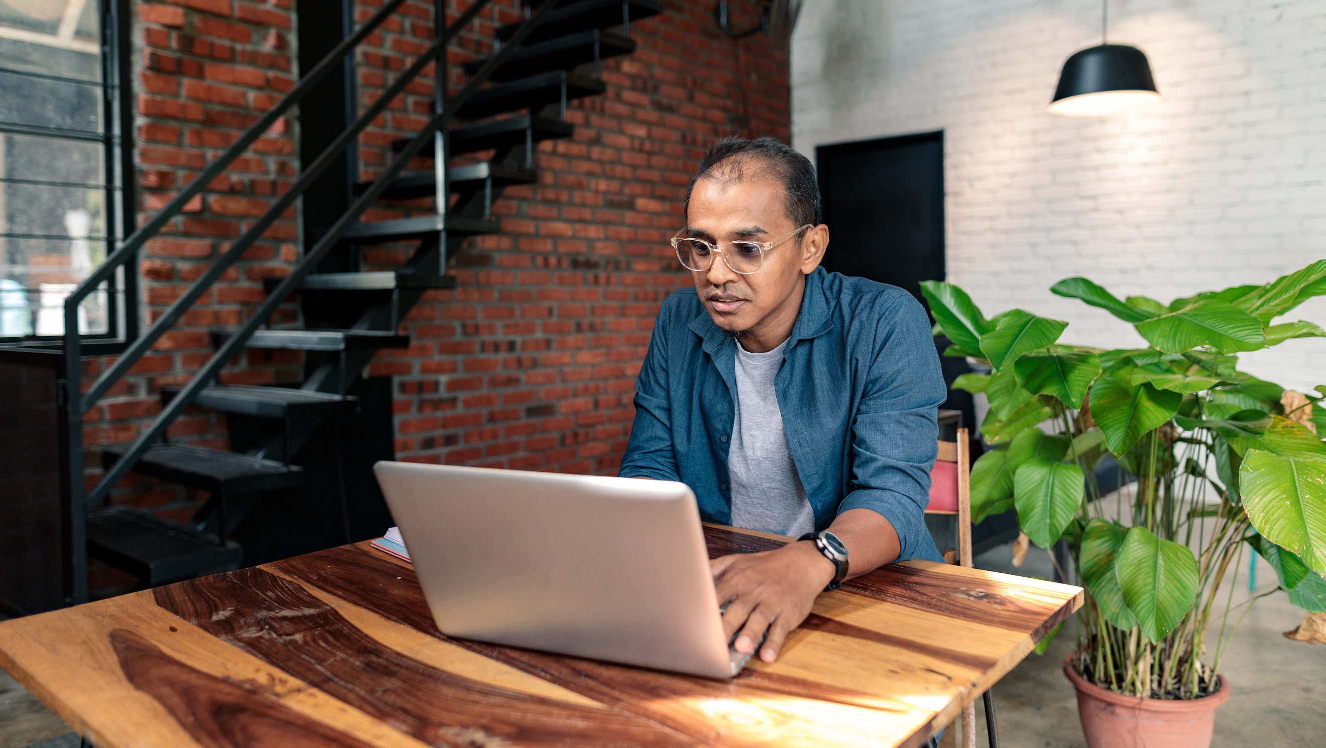 A man with glasses sits at a high top wooden table while working on his laptop.