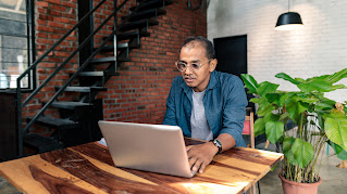 A man with glasses sits at a high top wooden table while working on his laptop.