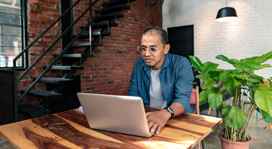 A man with glasses sits at a high top wooden table while working on his laptop.