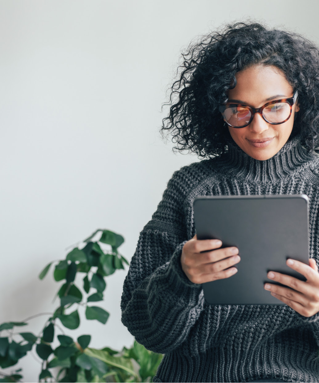 A young woman in a gray sweater looks at the screen of a tablet. She stands next to a sunny window, a plant is behind her.