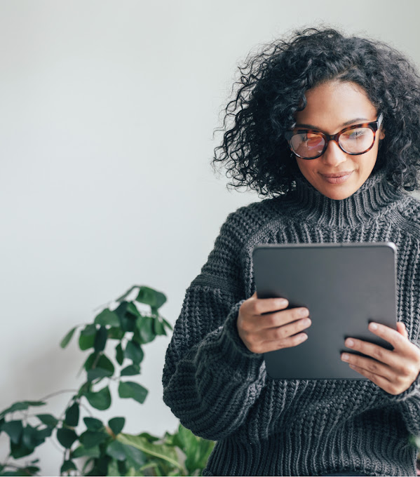 A young woman in a gray sweater looks at the screen of a tablet. She stands next to a sunny window, a plant is behind her.