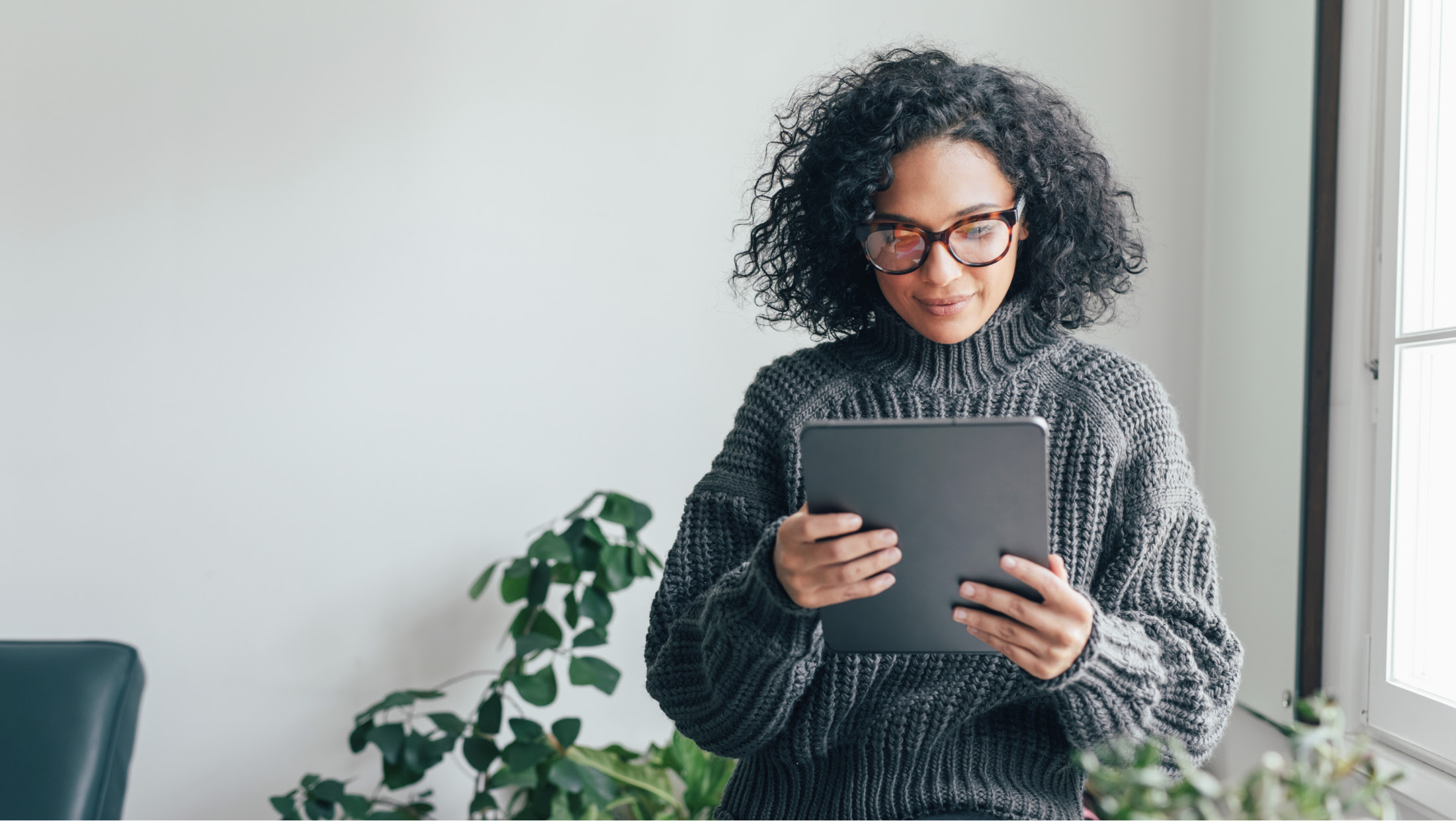 A young woman in a gray sweater looks at the screen of a tablet. She stands next to a sunny window, a plant is behind her.