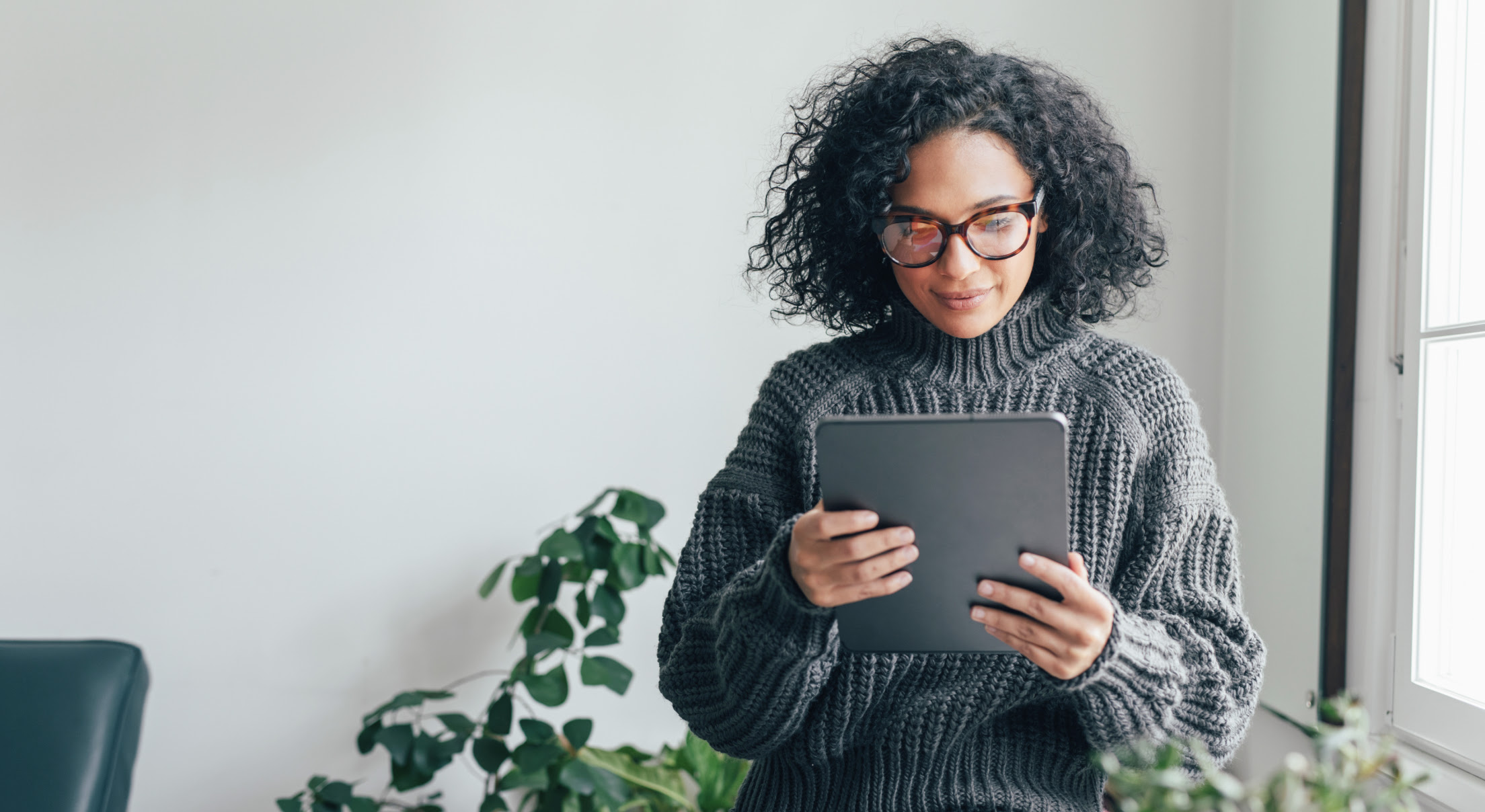 A young woman in a gray sweater looks at the screen of a tablet. She stands next to a sunny window, a plant is behind her.