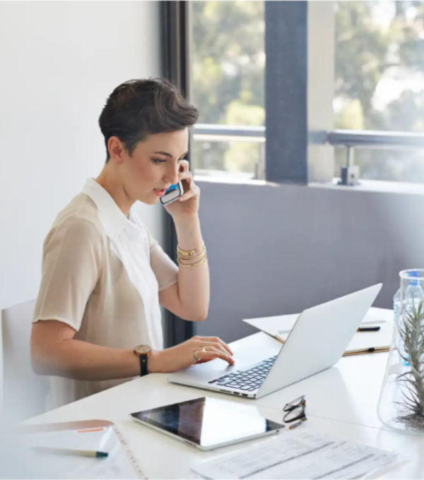 A short haired woman sits at a desk and talks on a smartphone with a laptop and tablet in front of her.