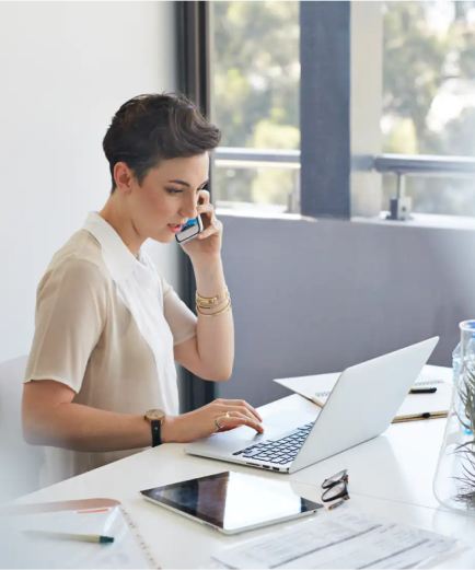 A short haired woman sits at a desk and talks on a smartphone with a laptop and tablet in front of her.