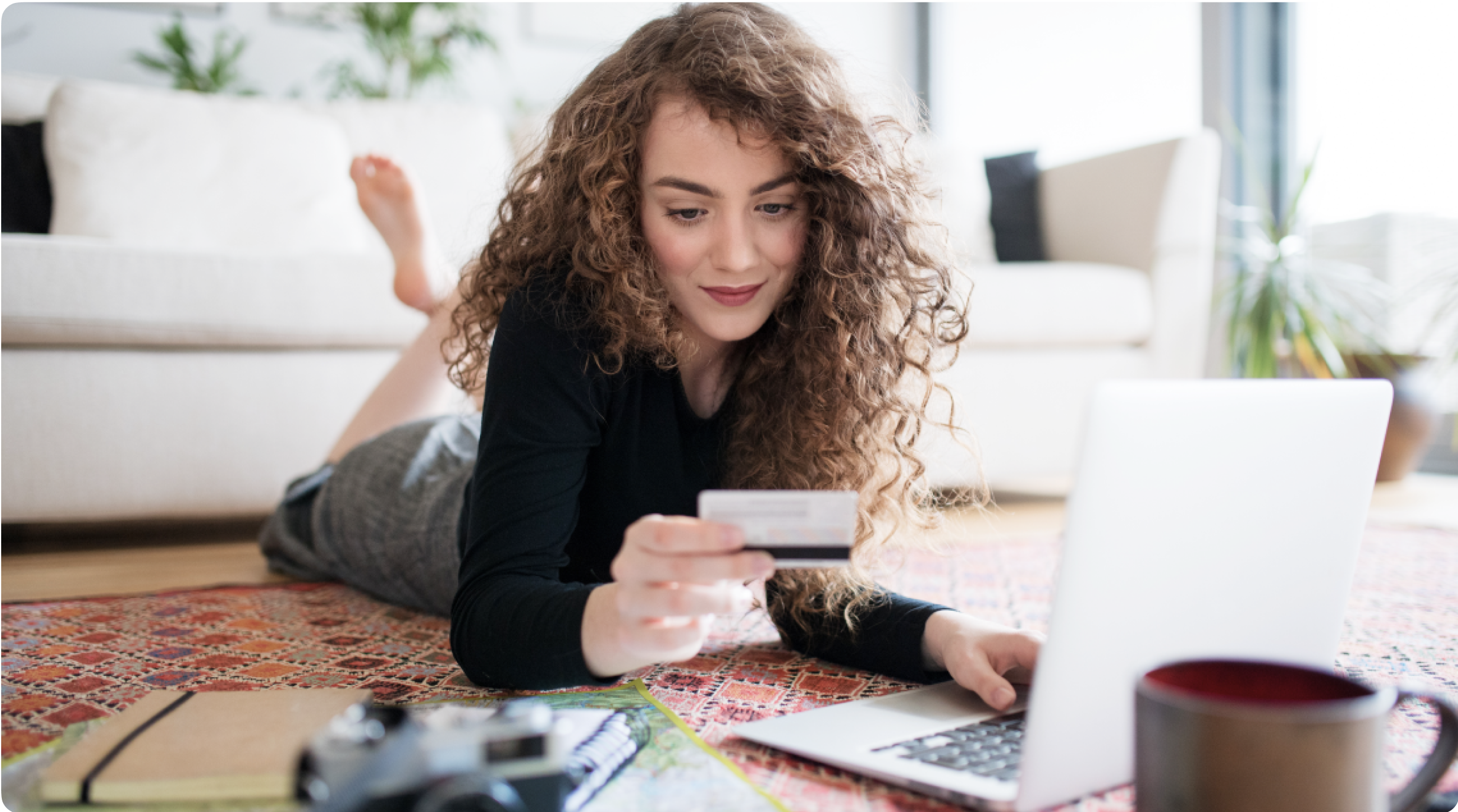 A woman with long, curly hair shops online from the floor. She types in a credit card number while laying on her stomach.