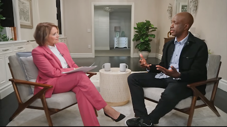 The journalist Katie Couric interviews a seated man; between them is a solid, round coffee table holding two coffee cups