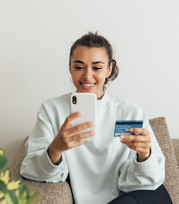 A young woman in a light blue top uses a credit card to complete a payment on her smartphone.