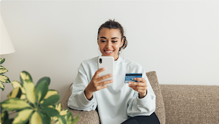 A young woman in a light blue top uses a credit card to complete a payment on her smartphone.