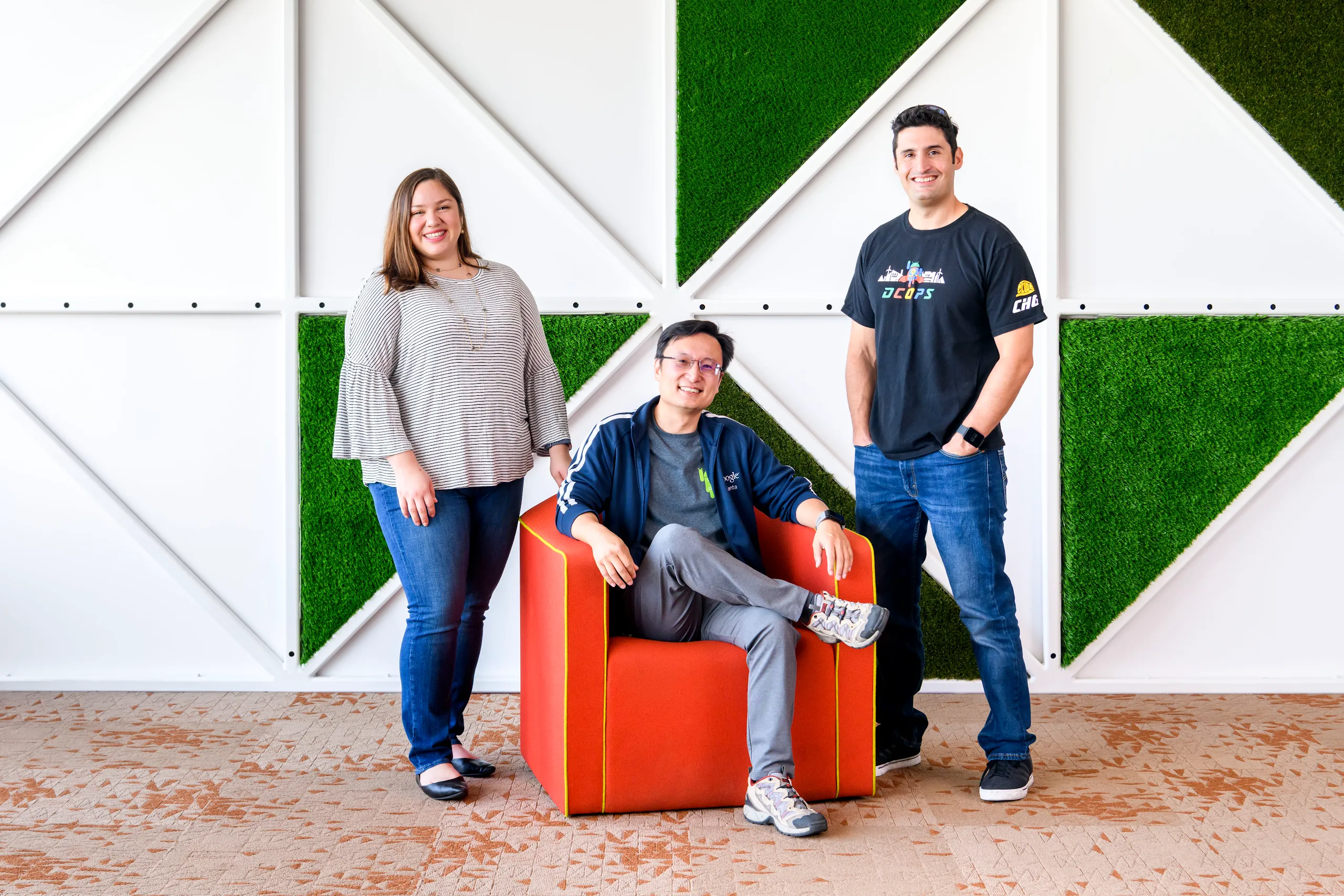 Photograph of three people smiling in front of a graphic wall in the background.