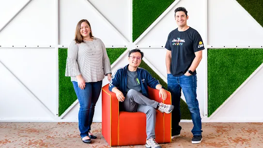 Photograph of three people smiling in front of a graphic wall in the background.