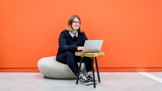 A photograph of a woman sitting down with a laptop looking at the camera.