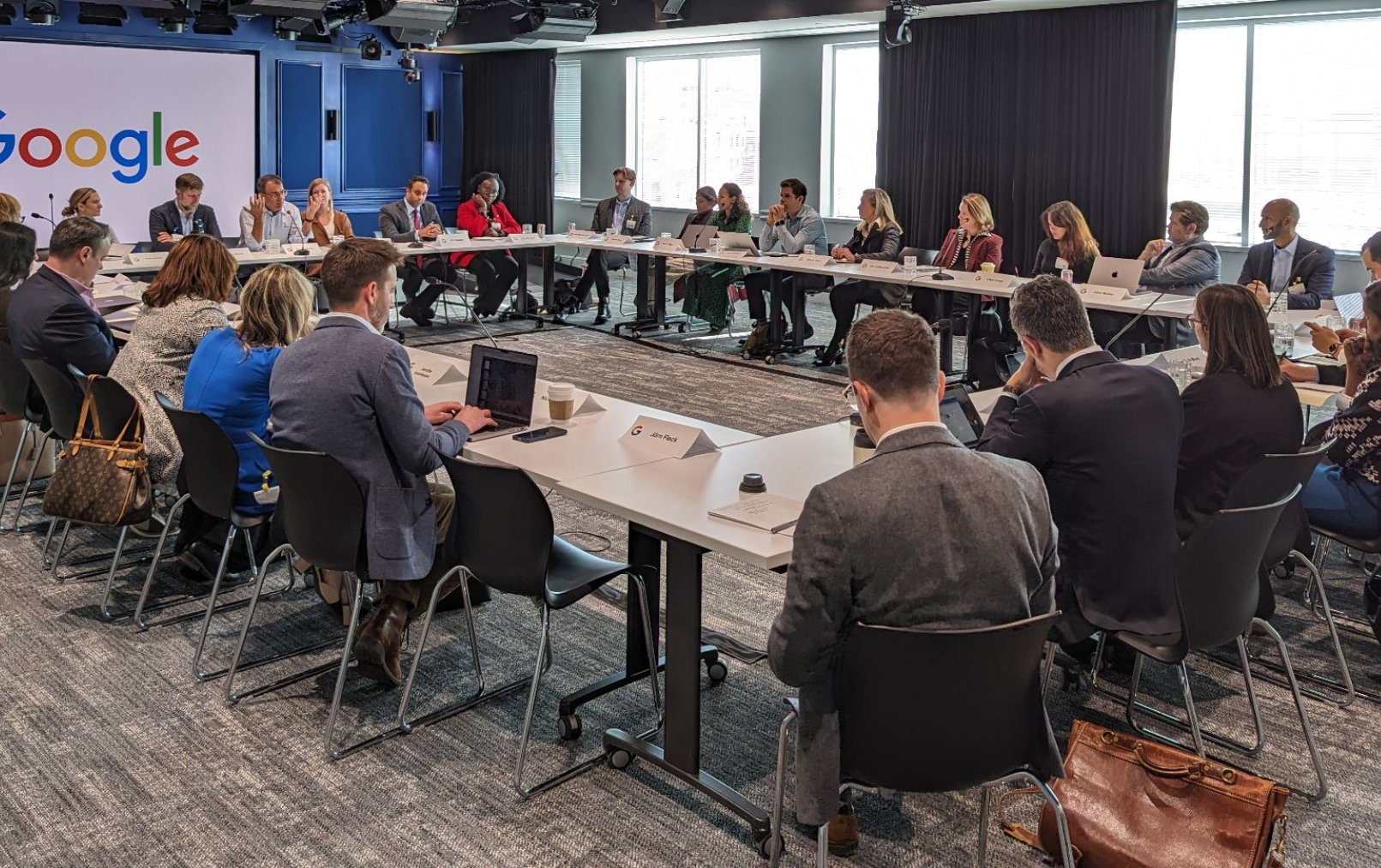 People sitting around tables in a square formation with a Google logo visible on a screen behind them