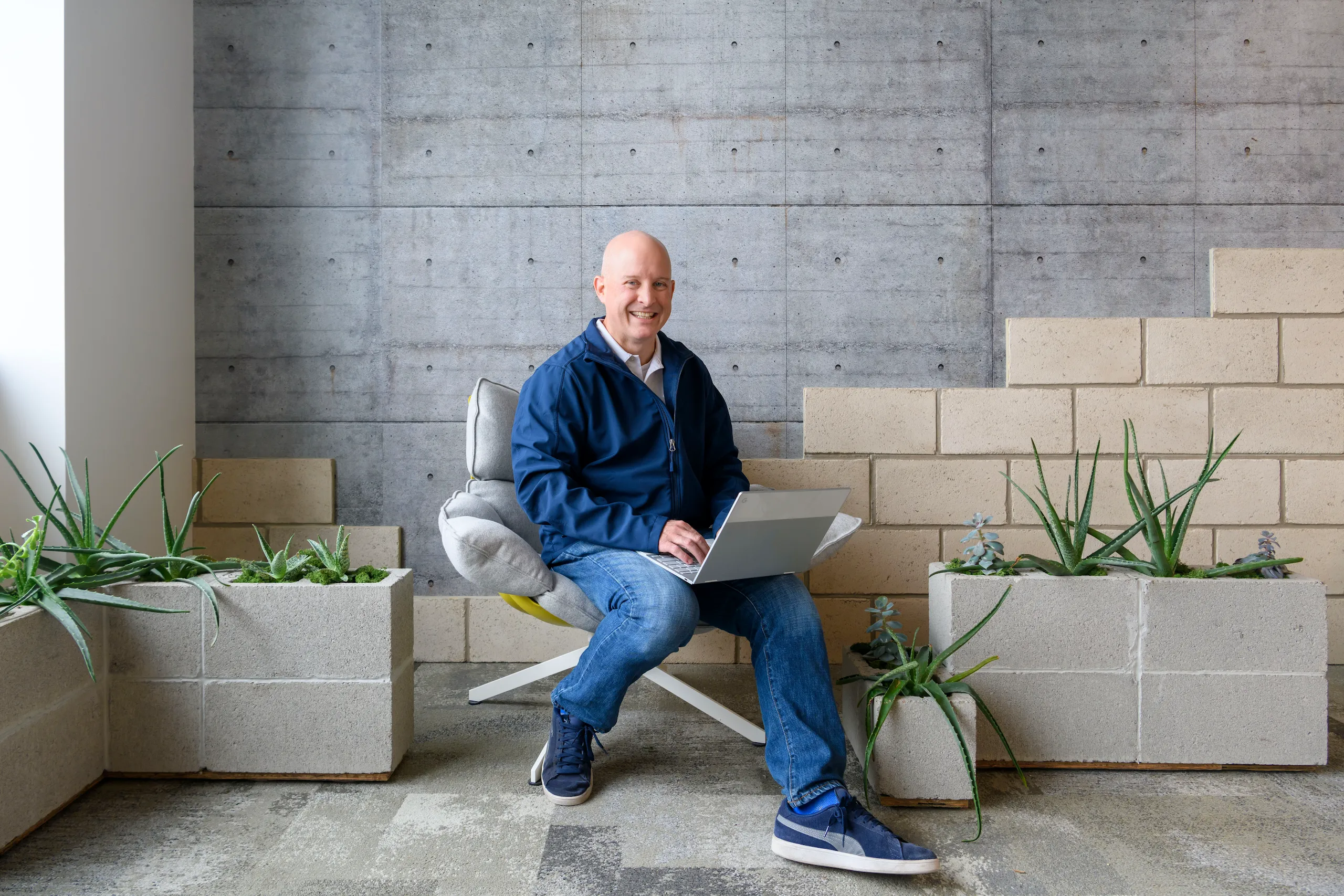 Photograph of a smiling man sitting down with a laptop surrounded by plants.