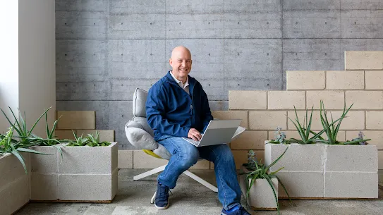 Photograph of a smiling man sitting down with a laptop surrounded by plants.