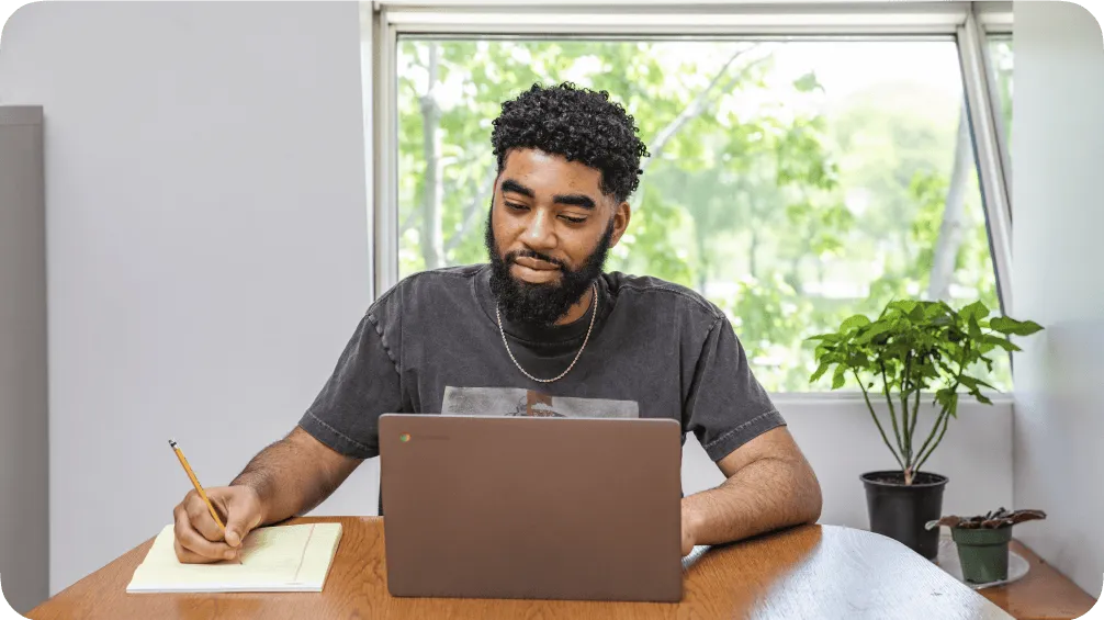 A man wearing a black t-shirt works on a laptop while taking notes in a legal pad