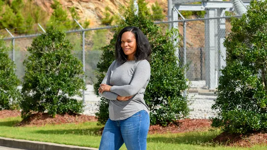 A photograph of a woman in a grey tshirt with arms folded standing outside looking at camera.