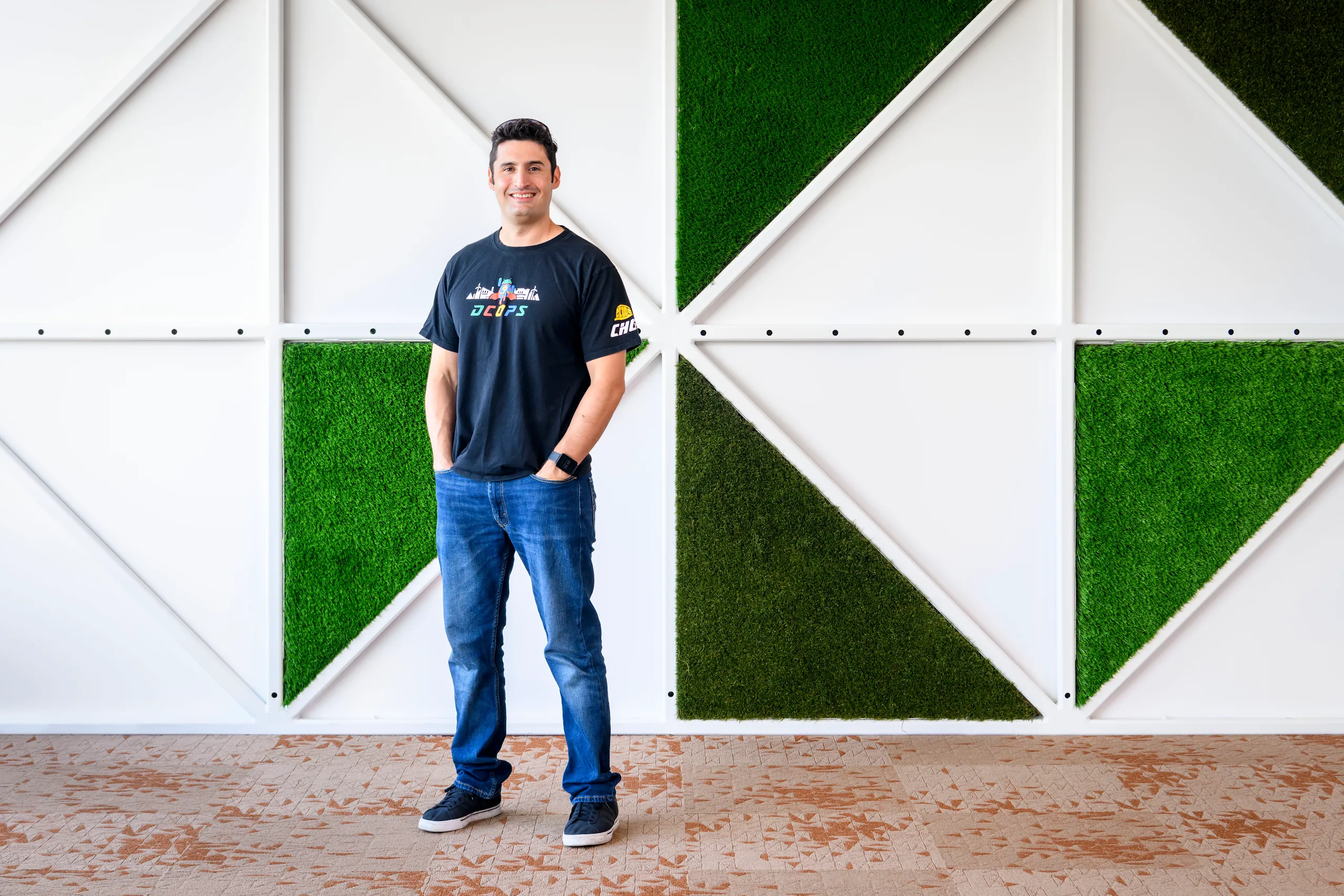 Photograph of a smiling man standing with hands in pocket in front of a graphic wall in the background.