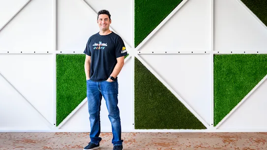 Photograph of a smiling man standing with hands in pocket in front of a graphic wall in the background.