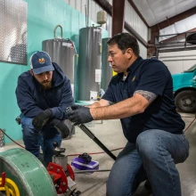 Two members of the A&A Plumbing team, wearing dark blue shirts and jeans, discuss a piece of equipment in a workshop. A truck is visible in the background.