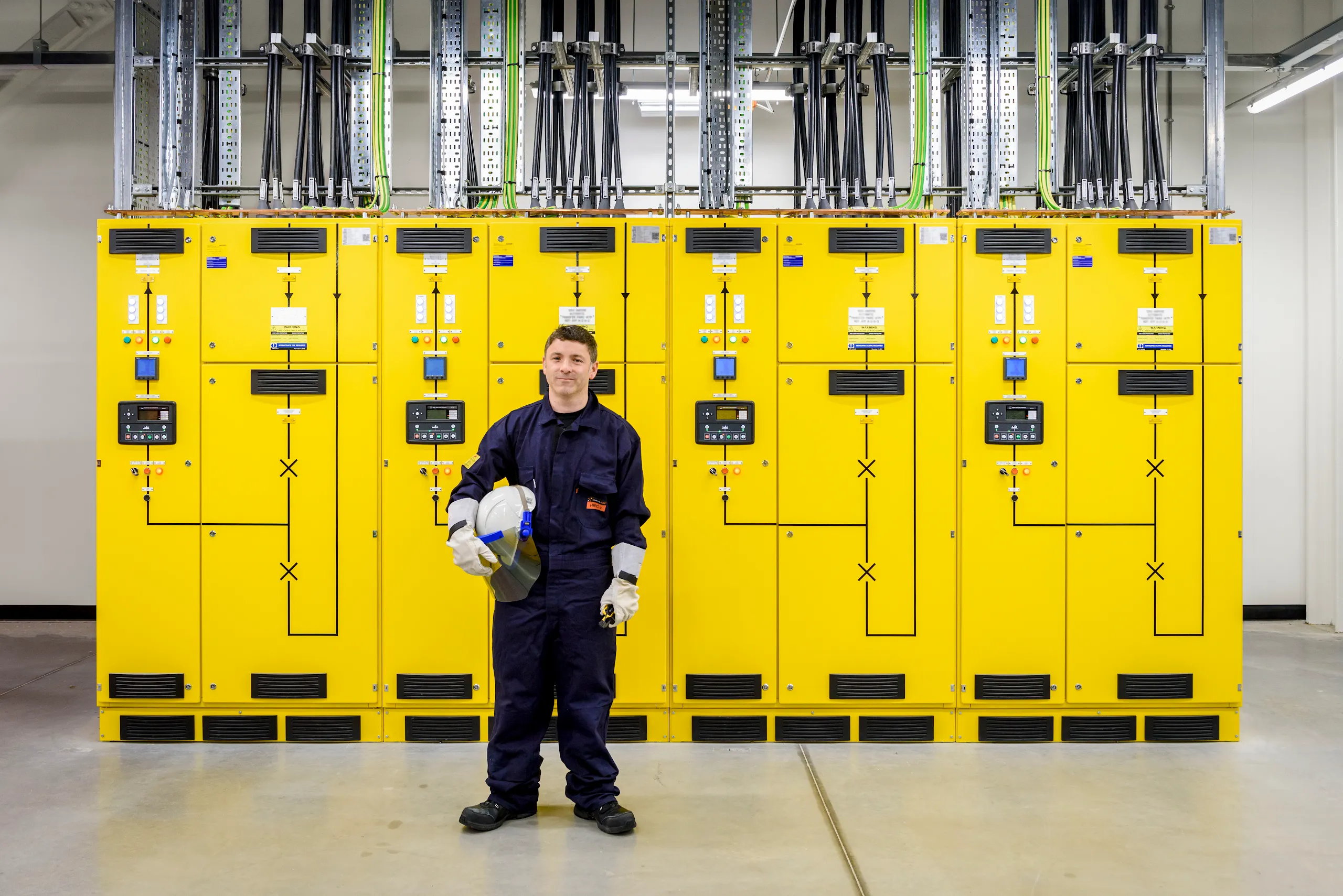 A photograph of a man standing in a facilities technician uniform in front of electrical systems.