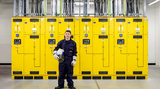 A photograph of a man standing in a facilities technician uniform in front of electrical systems.