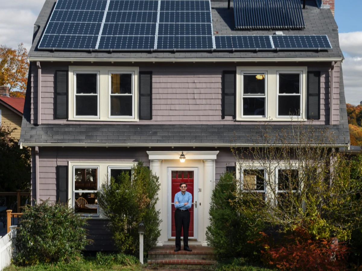A man stands in front of a quaint home with solar panels on the roof.