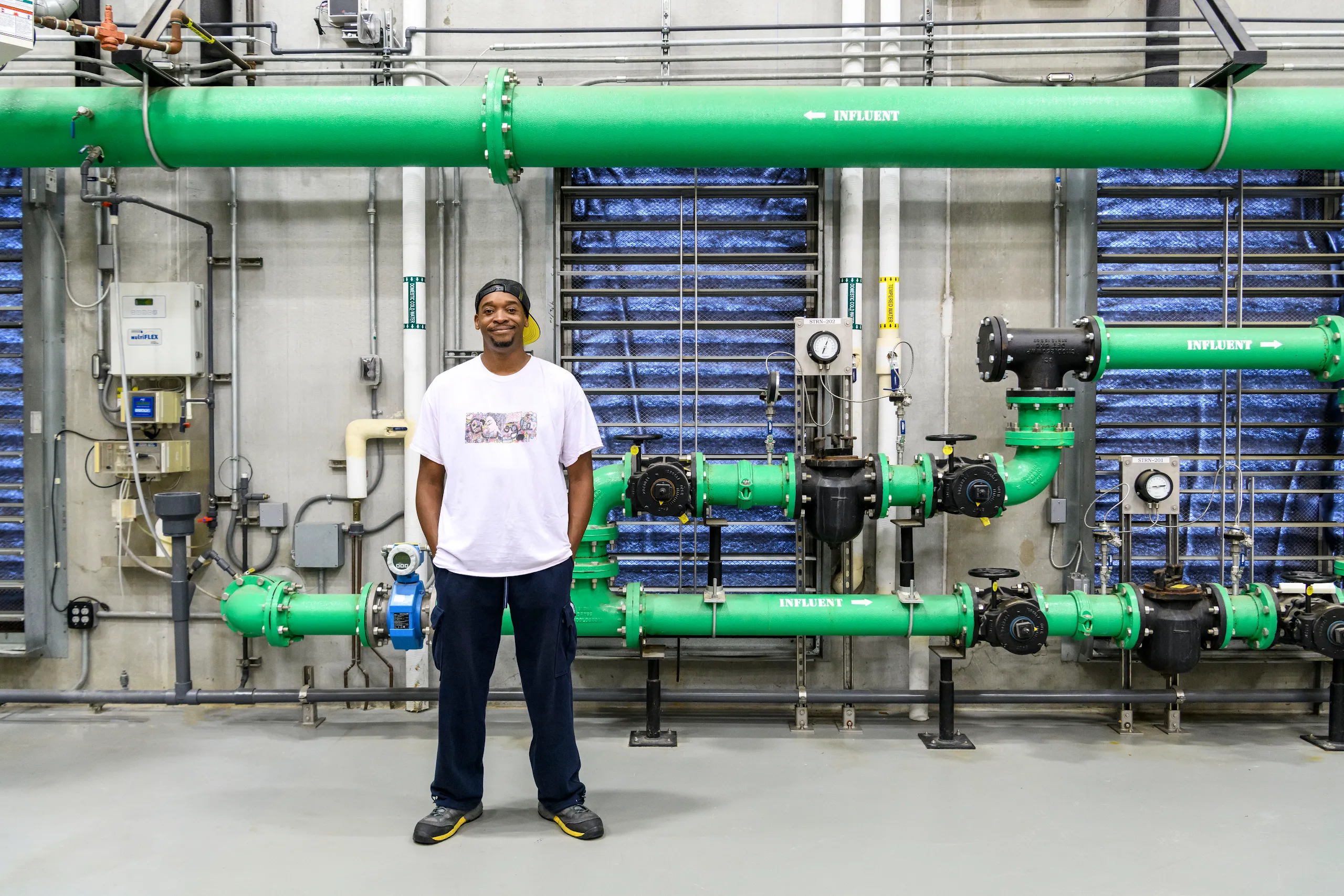 A full body photograph of a man wearing a tshirt with hands in pocket standing in front of colorful pipes in a Google facility.