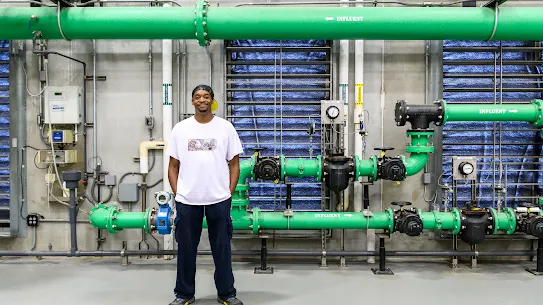 A full body photograph of a man wearing a tshirt with hands in pocket standing in front of colorful pipes in a Google facility.