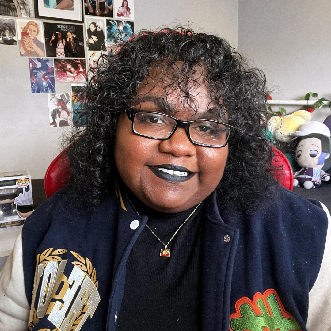 Photograph of Naarm woman wearing a navy blue and beige varsity jacket, black shirt, and glasses with short curly black hair sitting in a chair. 