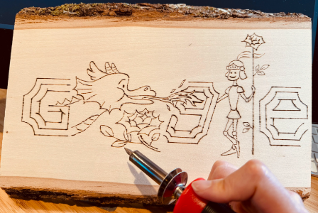 The unfinished Doodle sketched into a piece of wood with the artist's hand holding an engraving pen.