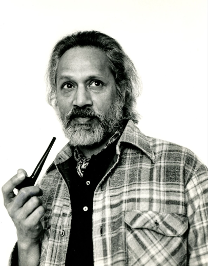 Black and White photo of Lancelot Ribeiro holding a pipe. He has medium toned skin with dark/gray hair, gray beard and wearing a plaid jacket with a black shirt under.