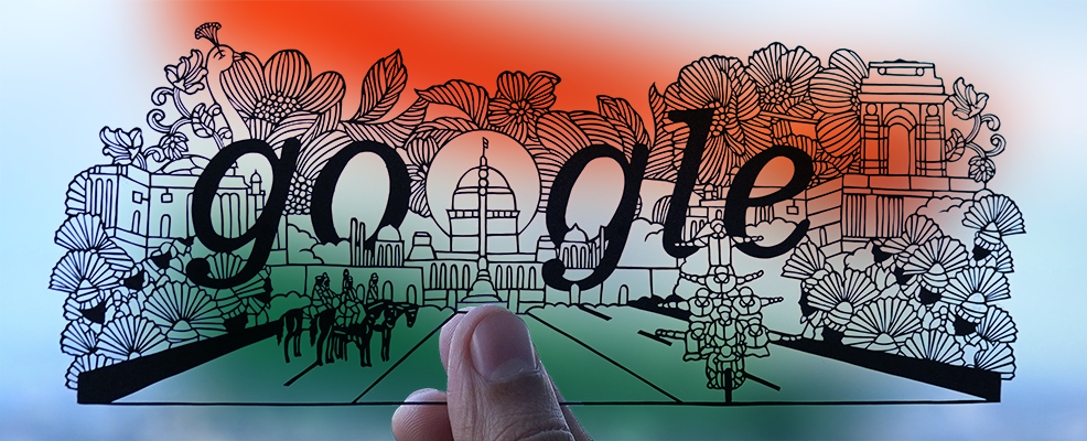 Photograph of an intricate paper cut-out of an Independance Day celebration scene in India held by a thumb and forefingers. The "GOOGLE" logo is positioned prominately in the background with a domed building standing in as the second "O". In the foreground, men on horseback are on the left and a stack of men riding a singular motocycle are on the right. Decorative fans and flowers surround the scene and a large bird and archway are tucked into the background. A blue sky with white clouds can be