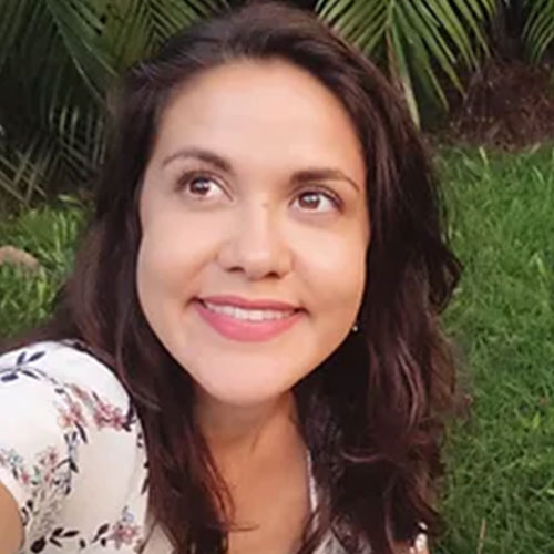 Headshot photograph of Samantha. Samantha has medium length, straight, brown hair and brown eyes. She is smiling and looking up at the sky. She is wearing a white top with pink flowers and is standing in font of greenery.