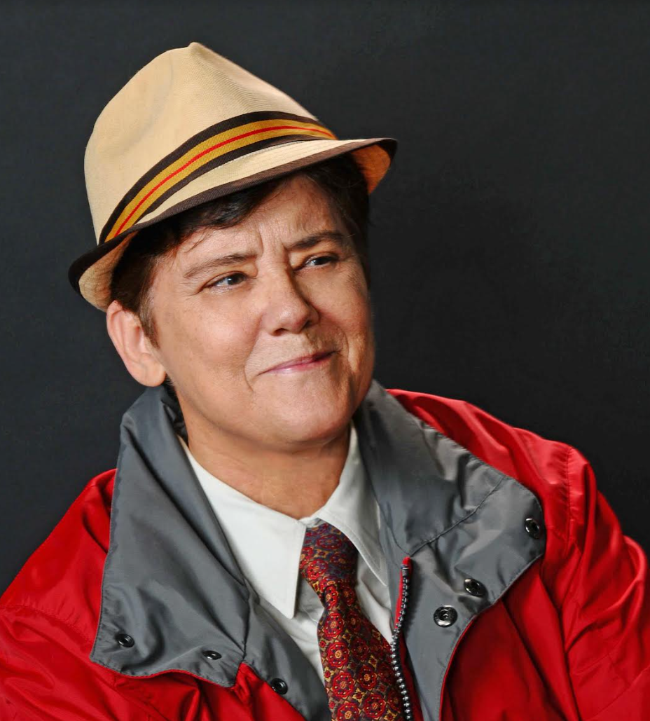 Photograph of Jeanne Córdova smiling, wearing a red jacket and beige hat.