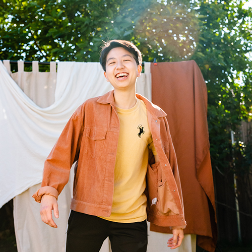 Photograph of a person with short black hair wearing an orange button up shirt smiling to camera in an outdoor setting.