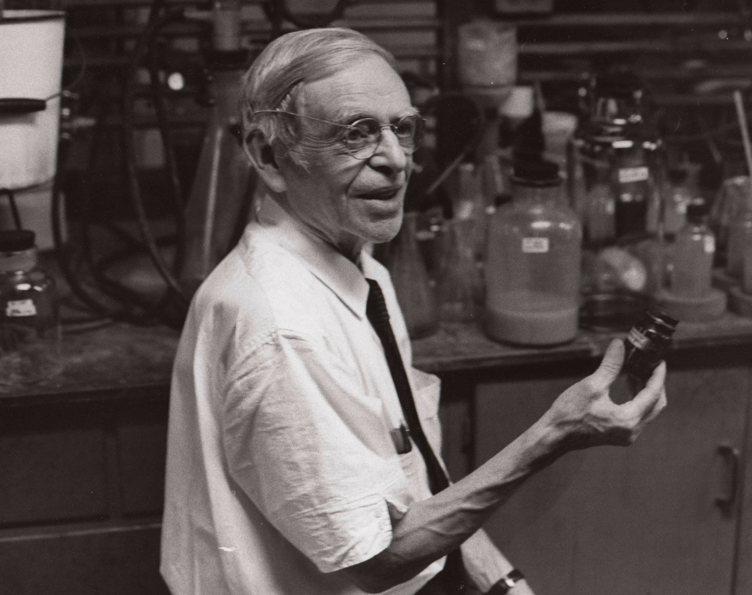 Black and white photograph of Casimir Funk holding a small bottle in a lab. He wears a white short sleeve shirt, a black tie, and round eye glasses.