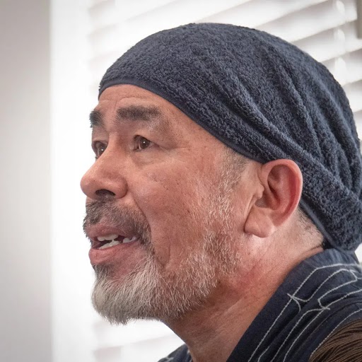 Photograph of a middle aged Asian man with facial hair looking off camera wearing piece of fabric on his head. 