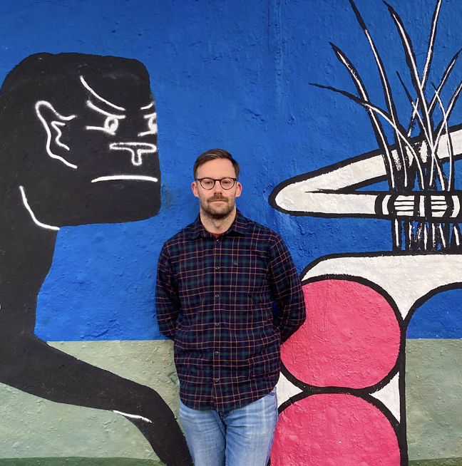 Photograph of a man standing in front of a colorful painted wall. He has facial hair, glasses, and is wearing a dark shirt.