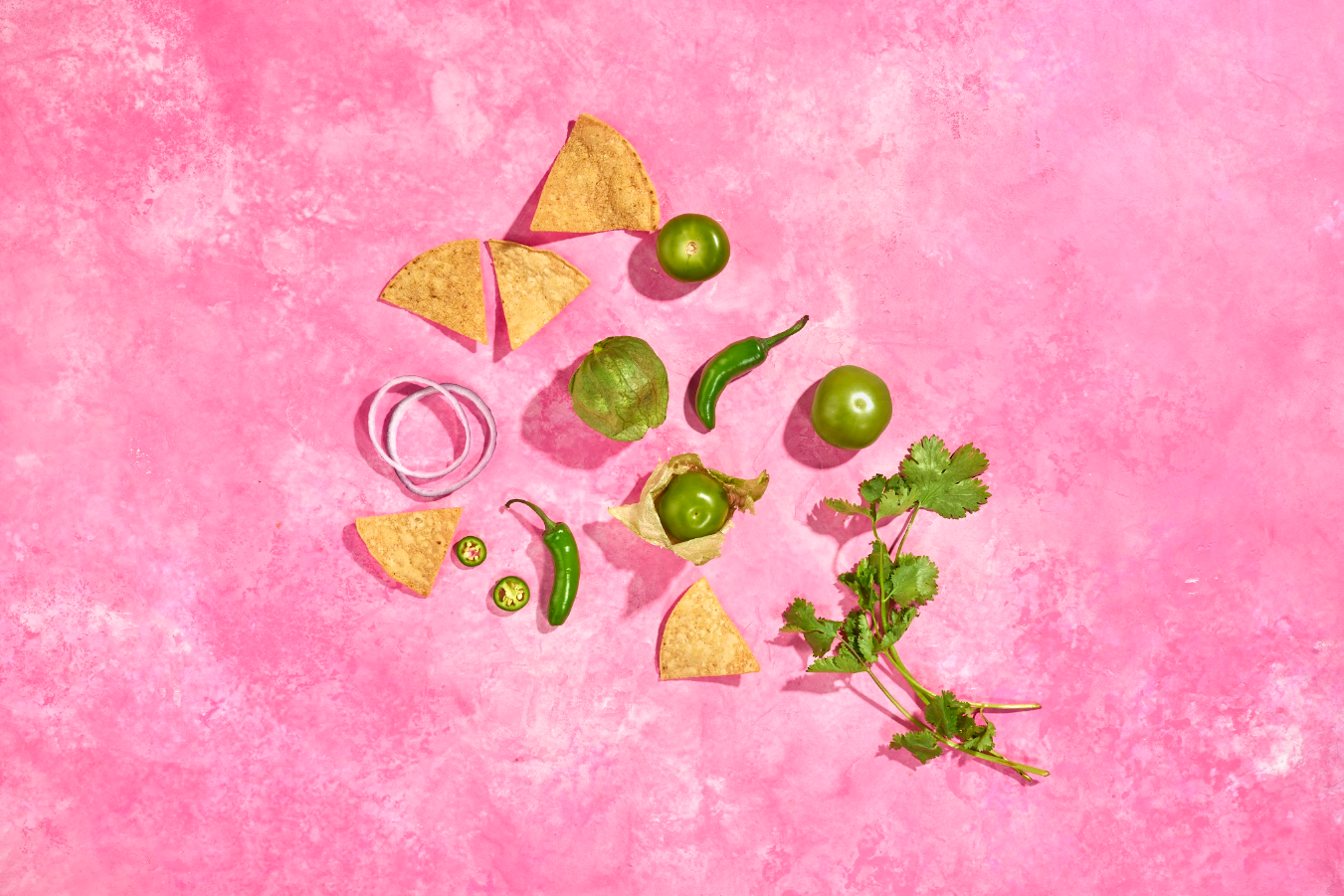 Image of the tortilla chips, cilantro, jalepeños and onions laid out on a pink background